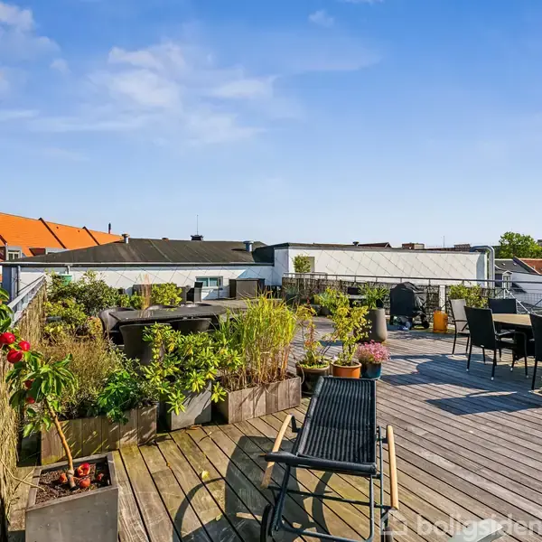 Terrasse med træbeklædt gulv og havemøbler omringet af blomsterkrukker. Lige op til røde tagsten på nabobygninger, under en klar blå himmel uden tekst.