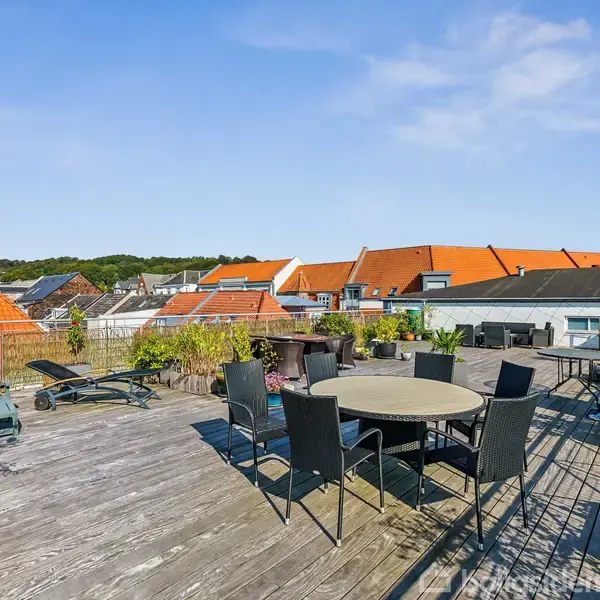 En stor træterrasse med havemøbler, herunder runde borde og stole, omgivet af plantepotter. Terrasse ligger på en tagtop med udsigt over hustage med orange tegl under en klar blå himmel.