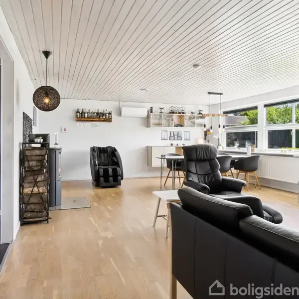 Stue med sorte lædersofaer står i et åbent rum. Et spisebord med stole er ved store vinduer. Lyse trægulve, hvidt loft, og moderne indretning skaber en lys og indbydende atmosfære.