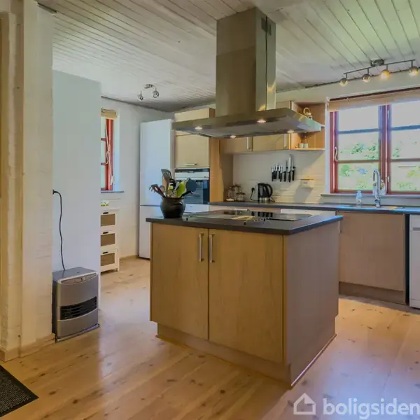 Køkken med træskabe står i rummeligt rum. Kogeø med emhætte i midten. Store vinduer i baggrunden giver naturligt lys. Vaskemaskine og diverse køkkenredskaber er synlige.