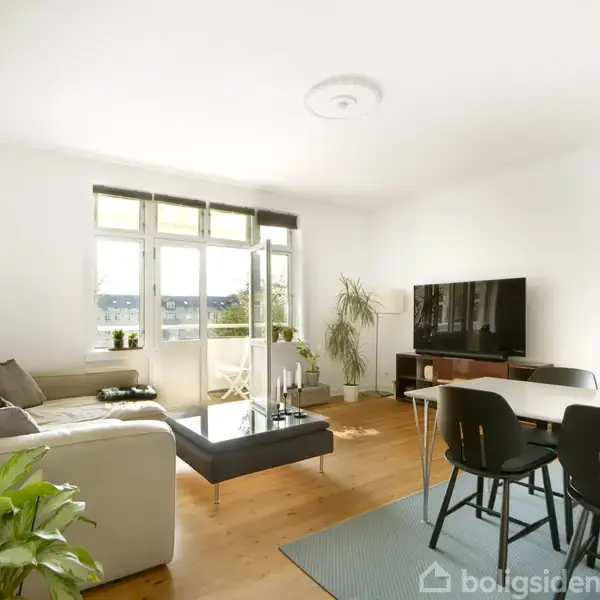I en lys stue står en L-formet sofa ved siden af et kaffebord. En stor fladskærm er placeret på et mediebord, omgivet af grønne planter og spisebord med stole.