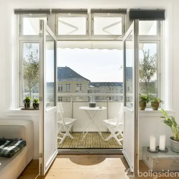 En balkon med et lille rundt bord og to stole åbner op fra en stue. Døre står på vid gab, og planter ses både indendørs og udendørs.
