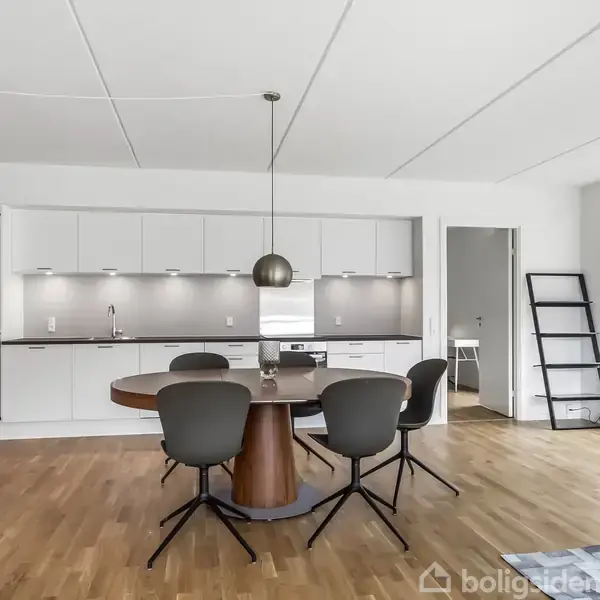 Et spisebord med fire stole står i et moderne køkken-alrum. Køkkenet har hvide skabe og grå backsplash, mens et stort vindue tilføjer naturligt lys. En stigehylde står til højre.