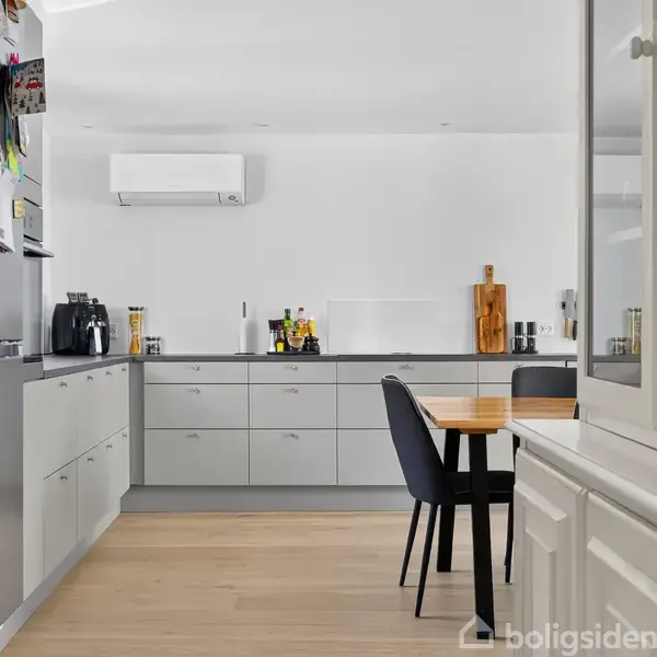 Køkken med grå skabe og en sort bordplade. En spisestol står ved et træbord. Køkkenudstyr ses på skabene, mens en airconditionenhed hænger på væggen.