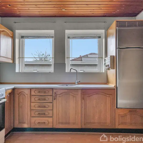 Køkken med trækøkkenskabe og stålkomfur, rustfri stål køleskab, og en vask under to vinduer. Træloft og hvide vægge skaber en lys og rummelig atmosfære.