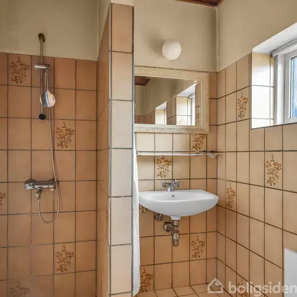 En bruser og håndvask monteret på væggen i et badeværelse med lysebrune fliser, der har et blomstermønster. Ved siden af er et lille vindue, og til venstre et blåt badeforhæng.