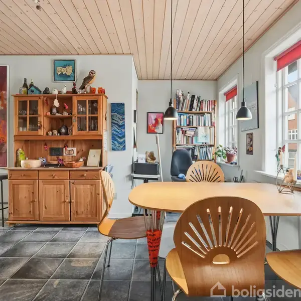 En lys spisestue med træbord og stole, indeholder en kreds af lamper. Træskænk fyldt med dekorationer. Store vinduer med røde rullegardiner giver udsigt til nabolaget.