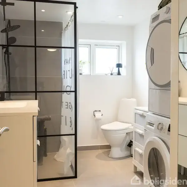 Badeværelse med bruseniche adskilt af glasvæg. Vaskemaskine og tørretumbler er stablet ved siden af et rundt spejl. Toilet ved vindue i lys, moderne indretning.