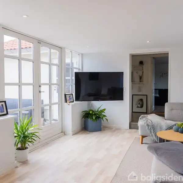 Stue med en grå sofa, en tv-skærm på væggen og et sofabord i et lyst rum. Store vinduer med planter i vindueskarmen skaber en lys atmosfære.