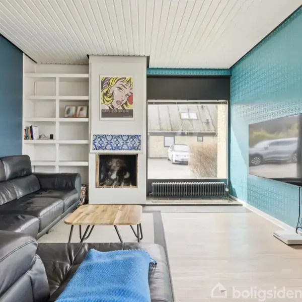 Sort lædersofa står i en moderne stue med blå accentvægge. Et sofabord står foran sofaen. En pejs og en vægmonteret tv-skærm er i rummet. Maleri over pejsen.
