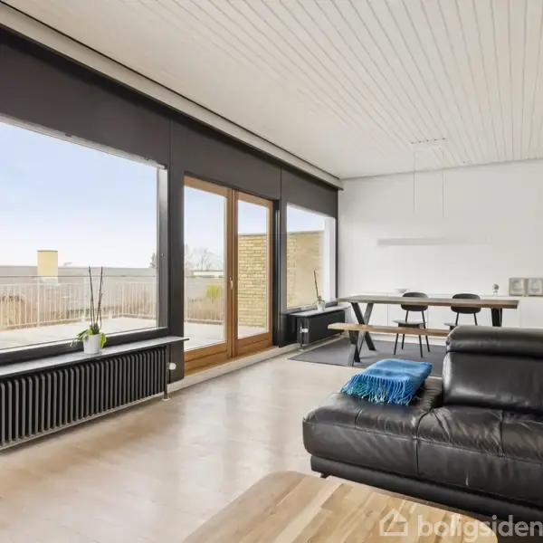 Sofa i sort læder står i stue med trægulv. Ved siden af er spisebord med stole. Store vinduer giver udsigt til terrasse med planter udenfor.
