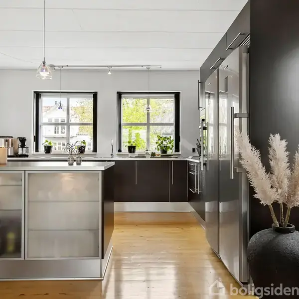 Et moderne køkken har et stort gråt køleskab og en ø med glaslåger. Det er lyst med vinduer, planter og hængende lamper, skaber en indbydende atmosfære.