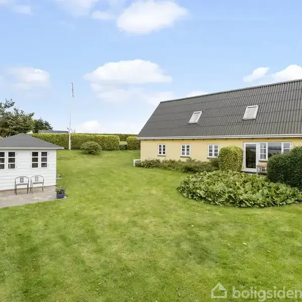 Et gult hus med sort tag står i en velplejet have med en hvid haveskur til venstre. Omkringliggende buske og træer skaber en landlig atmosfære under en blå himmel.