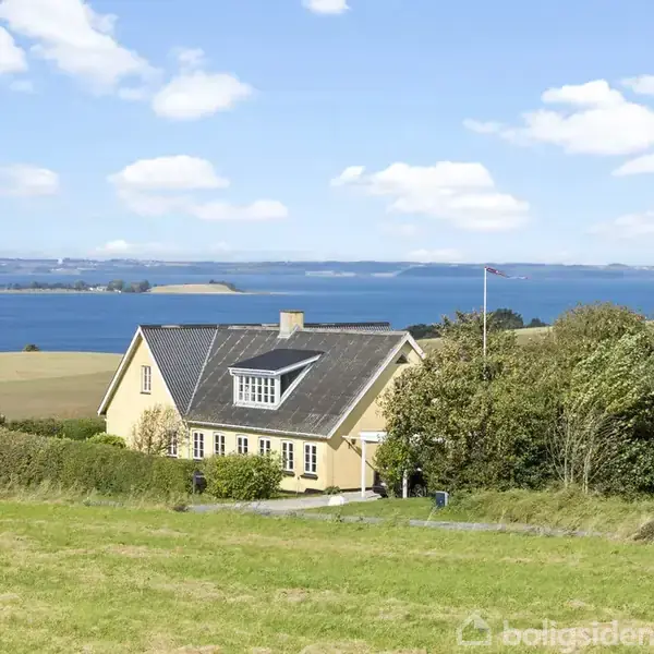 Et gult hus står på en grøn bakke med udsigt over en blå fjord. Der er spredte træer omkring huset, og himlen er klar med nogle få skyer.