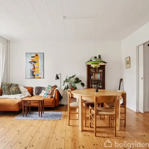 En stue med en brun lædersofa placeret ved et stort vindue. Et lille bord står foran sofaen, og et spisebord med stole er i rummet. En bogreol er i baggrunden. Væggen har et farverigt maleri.