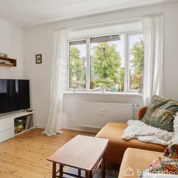 En stue med en fladskærms-tv stående på et hvidt tv-møbel. En orange sofa med tæpper og puder er placeret ved et vindue med udsigt til træer udenfor.