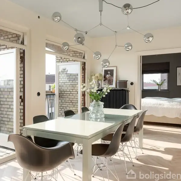 Et moderne spisestuemiljø med hvidt spisebord og brune stole. Bordet er pyntet med en glasvase med blomster. Store vinduer og åbne døre leder ud til en balkon. I baggrunden ses et soveværelse.