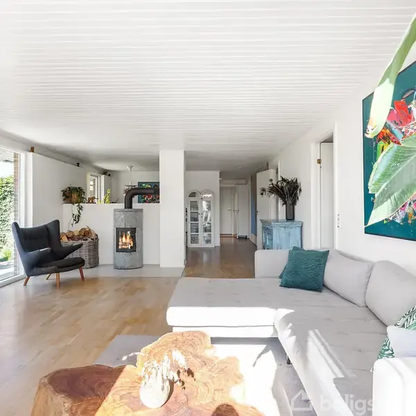 Stue med lys grå hjørnesofa foran et stort farverigt maleri. I baggrunden ses en pejs, kurvestol og store vinduer, der giver naturligt lys. Trægulv og moderne indretning.