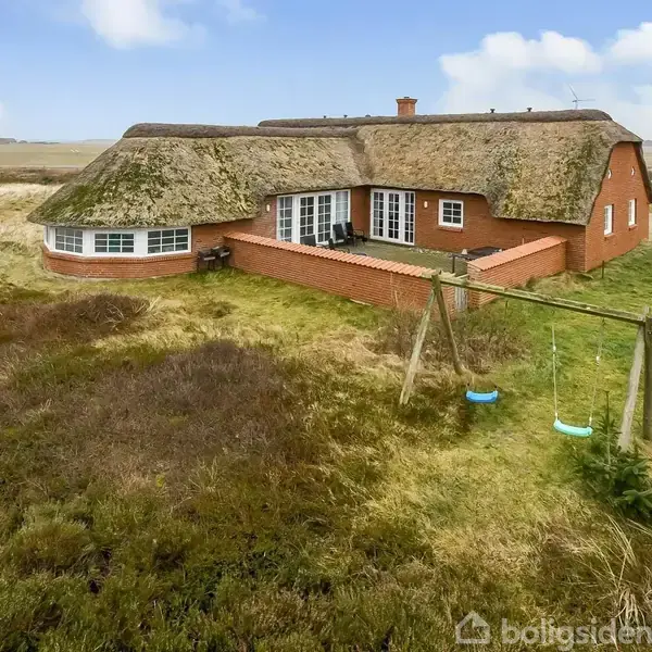 Stråtækt hus ligger i naturligt landskab med terrasse ved siden af det. Gyngestativ med to gynger står i græsset nær huset, omgivet af lyng og enebærbuske.