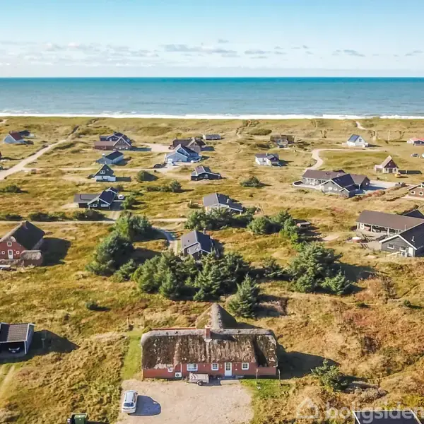 Hus med stråtag og røde murstensvægge står i forgrunden omgivet af græs og træer. Små huse spredt over klitlandskab i nærheden af en bred, sandstrand under en blå, klar skyfri himmel.
