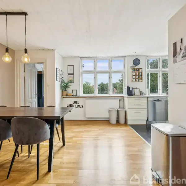 Spisebord med stole står i et lyst køkken-alrum med trægulv. Tre pendellamper hænger over bordet. Bagved er der hvide køkkenskabe, vinduer og teksten "LULU LARA LINUS".