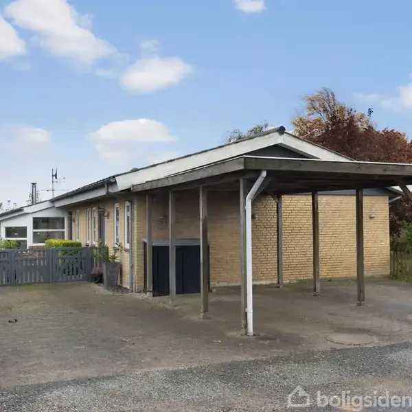 En murstensvilla står stille med en tilhørende carport i en forstadshave med en hæk omkring. Himlen er delvist skyet, og det er dagtimer.