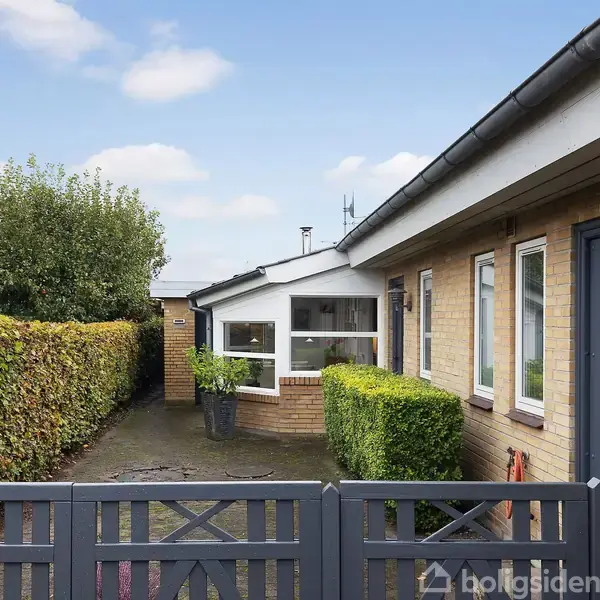 Et hus med gule murstensvægge og en blå dør står ved siden af en havegang. En bøgehæk danner en grøn grænse, mens en stor busk ses i baggrunden under en blå himmel.