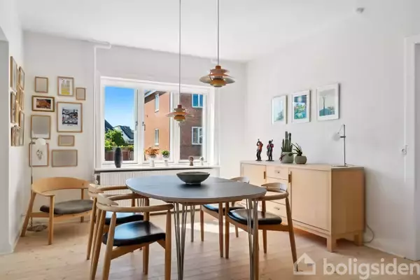 Spisebord omgivet af træstole står i et lyst rum. To lamper hænger over bordet. Vindue viser lejlighedsbygning. På væggene ses billeder og reoler med dekorationer.