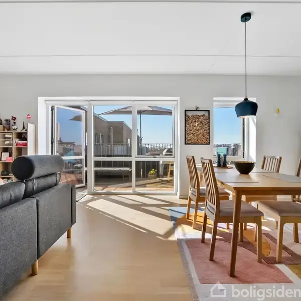 Stue med spisebord og stole, der står ved et stort vindue med udsigt til balkon og blå himmel. I baggrunden ses sofa, reoler med bøger og dekorative genstande.