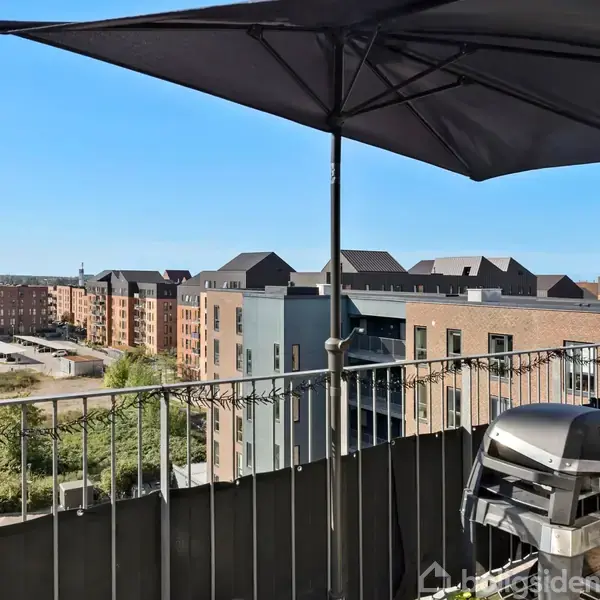 En altan med en grill og parasol. Udsigt over byens tage og boligblokke i solrigt vejr giver en afslappet og moderne atmosfære.