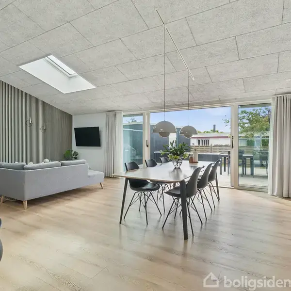 Moderne stue med spisebord og stole i centrum, omgivet af sofa og TV. Store vinduer giver udsigt til terrasse og have. Enkel indretning med lyse trægulve og grønne planter.