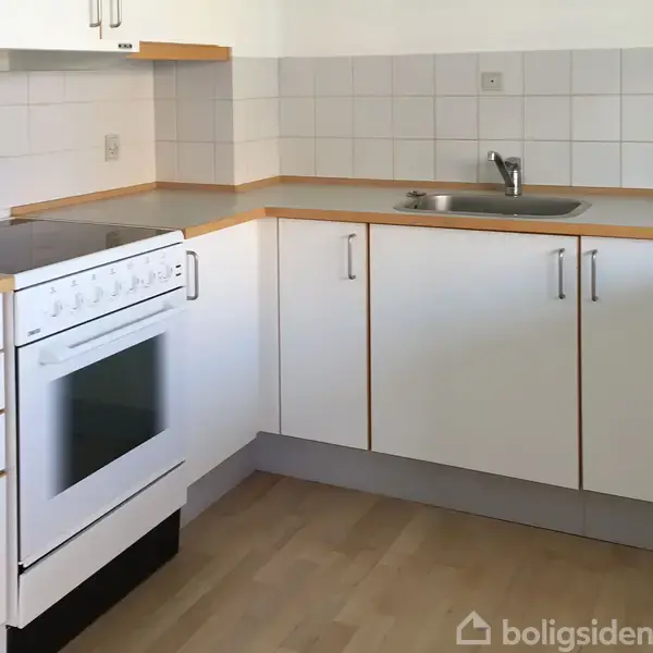 Komfur med hvid ovn i hjørnekøkken, omgivet af hvide skabe, lys trækant og gråt flisebagvæg. Håndvask til højre med enkeltstående vandhane, placeret på lyst trægulv.