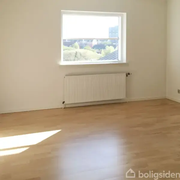 Et tomt rum med træparketgulv har et stort vindue, der lukker naturligt lys ind. En radiator er monteret under vinduet, og væggene er hvide.