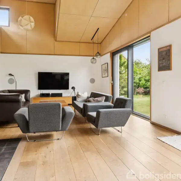 En moderne stue har en sort fladskærm monteret på en hvid væg, omgivet af to grå lænestole og en sofa. Store vinduer giver udsigt til en grøn have.