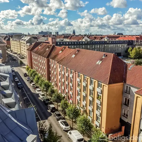 Række af murstensbygninger med røde tegltage, der strækker sig langs en træbevokset byvej, mens biler er parkeret på gaden. Solrigt vejr med spredte skyer over byens hustage.