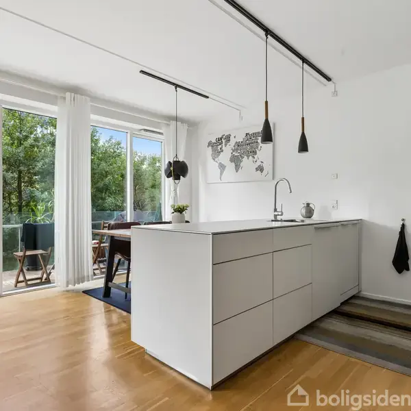 Et moderne køkken-alrum med en hvid køkkenø og hængende lamper. Gennem en glasskydedør ses en balkon med siddepladser og grøn udsigt. Verdenskort på væggen.