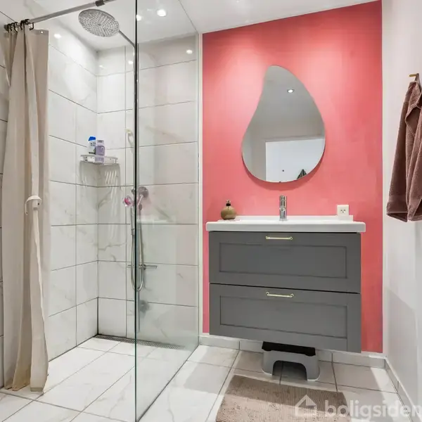 Brusekabine med glasvæg står ved siden af et gråt badeværelsesskab med spejl og håndvask. Pink væg i baggrunden, håndklæder hænger til højre.