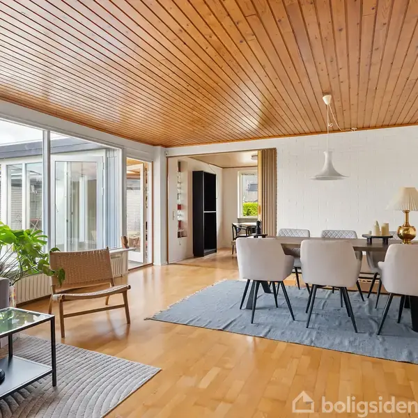 Stue med lyse træmøbler og sofa, sollys strømmer gennem store vinduer, planter pyntet ved siden af sofaen, spisebord med stole på gråt tæppe, maleri på væggen.