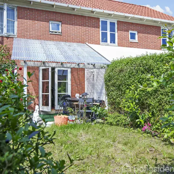 Røde murstensrækkehus med terrasse dækket af gennemsigtigt tag. Havemøbler står på terrassen. Foran er en velplejet have med buske og blomster i et roligt boligkvarter.