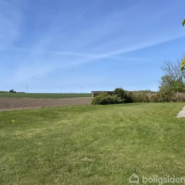 Græsplæne ligger ved siden af en murstensbygning. Himlen er klar med få skyer, og i baggrunden ses en mark og nogle buske.