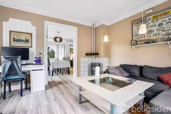 En stue med en grå sofa og glasbord foran en brændeovn. En skriftbord med computer er i hjørnet. Spisebord ses gennem en døråbning. Nummerplader hænger på væggen.