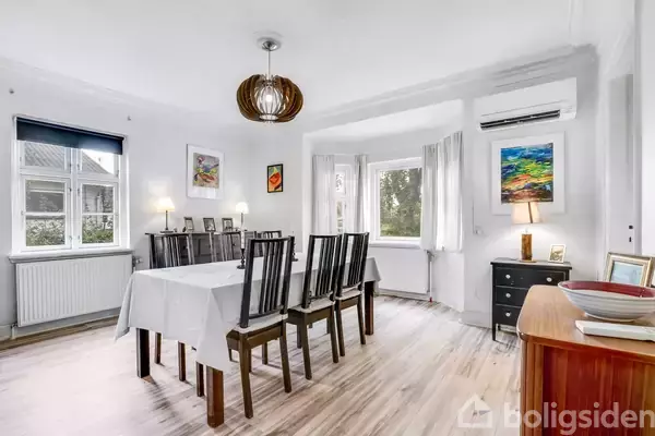 Spisebord dækket med hvid dug står i lys spisestue med trægulv. Seks stole omgiver bordet. Rummet er dekoreret med malerier, lamper og vinduer med udsigt til grønne træer.