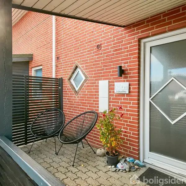 To stole står foran en rød murstensvæg. En glasdør med et diagonalt mønster er til højre. En lille plante står mellem stolene og døren. En lille syre på væggen siger "Velkommen til Villa Spangsbjerg".