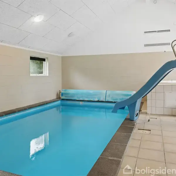 Indendørs swimmingpool med klar blåt vand og en blå rutsjebane på højre side. Rummet har lyst loft og vægge med to små vinduer. Ingen tekst til stede.