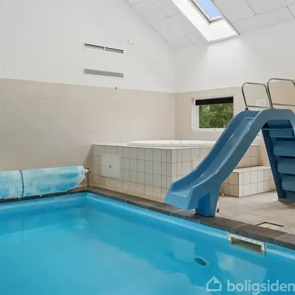 Indendørs swimmingpool med en blå vandrutsjebane fører ned i vandet. I baggrunden ses et spabad og en have udenfor gennem en glasdør.