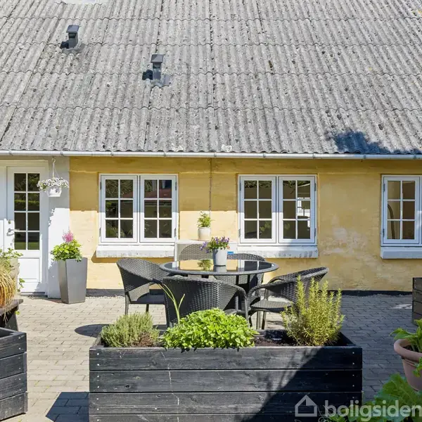 Terrasse med sort møbelsæt er placeret foran et gulmalet hus med hvide vinduer. Blomsterkasser omgiver området, der er belagt med fliser, hvor solen skinner.