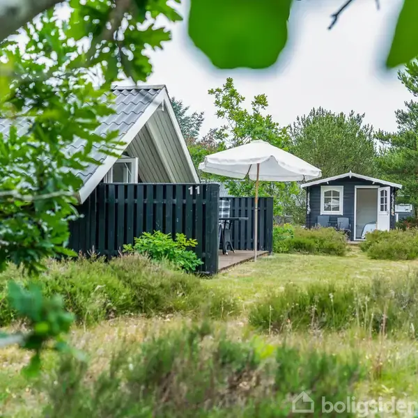 To små huse med mørke træfacader står i en grøn have. Et bord med parasol står på terrassen. Tallene "11" vises på hegnet.