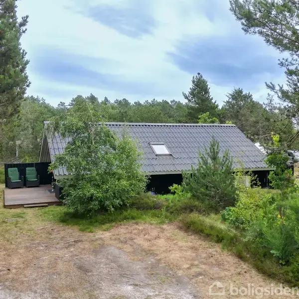 En sortmalet træhytte med gråt tag står stille i et skovområde, omgivet af træer og græs. Terrasse med havemøbler ses til venstre.