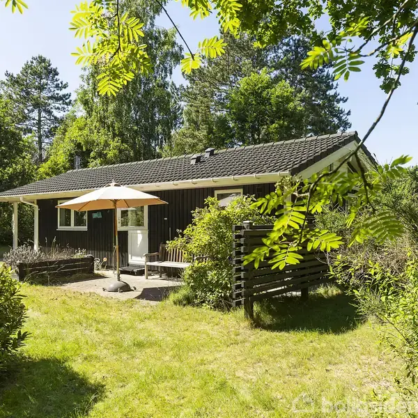 Sommerhus står stille i en grønt have, omgivet af træer og buske. En terrasse med parasol og stole skaber et hyggeligt opholdsområde foran huset.