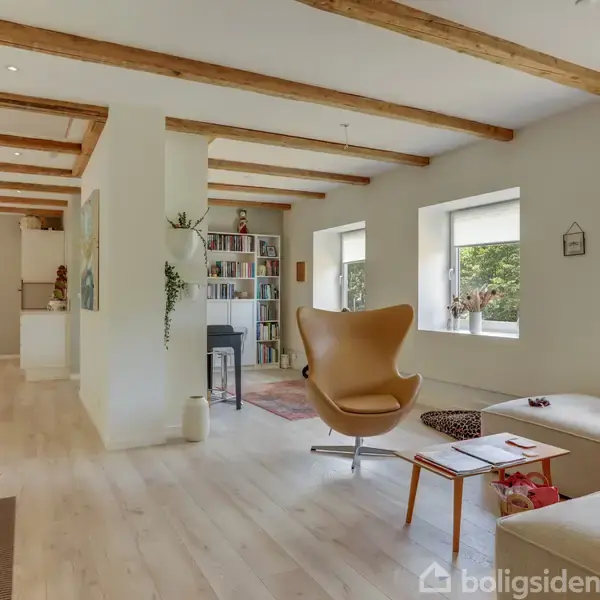 Et lyst opholdsrum med parketgulv, en lænestol og en sofa. Rummet har synlige loftsbjælker og store vinduer, der giver udsigt til grønne omgivelser.